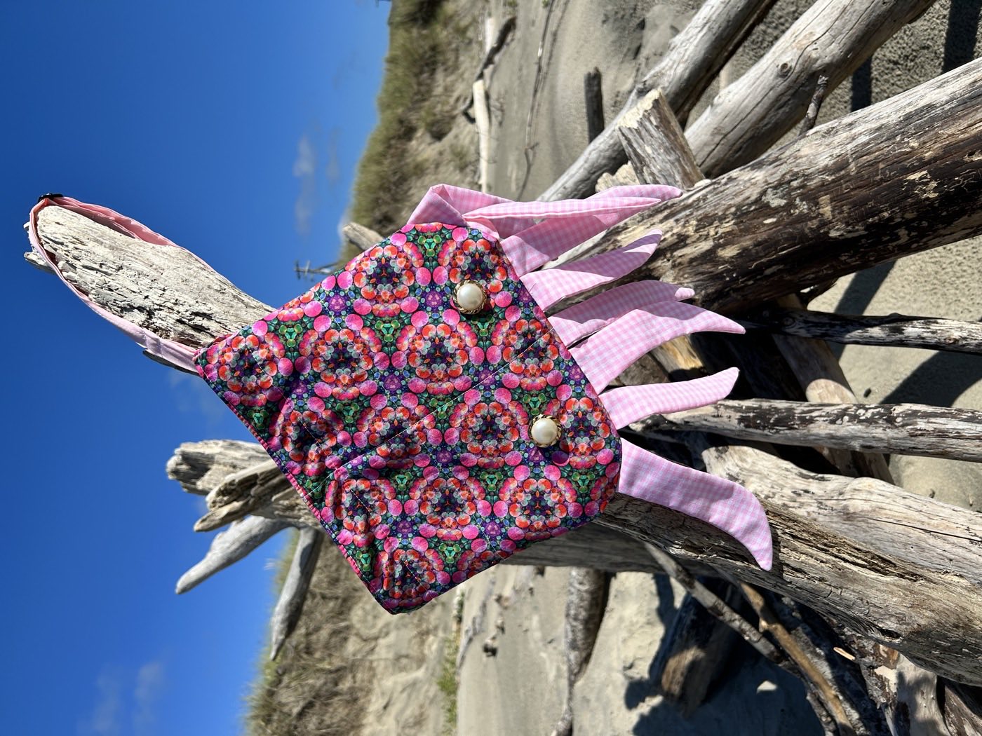 A small bag in a kaleidoscopic pink and green print, trimmed with pink gingham fringe and two pearl buttons, propped on driftwood under a blue sky.