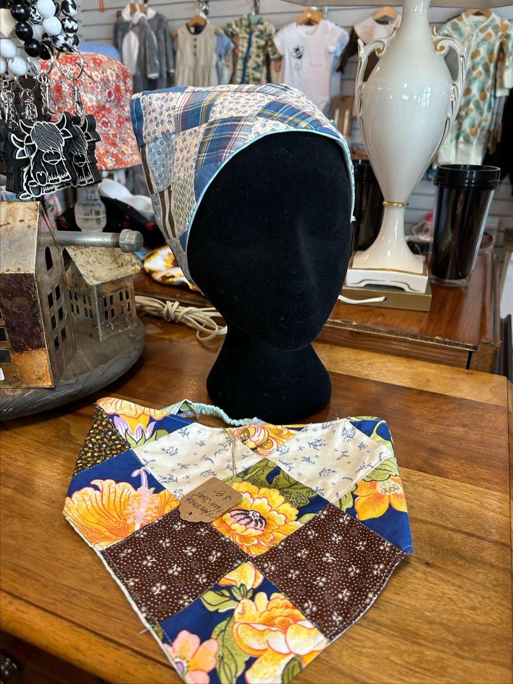 A blue plaid patchwork head scarf on a black mannequin head, with a brown and yellow floral patchwork bandana laid on the table in front.