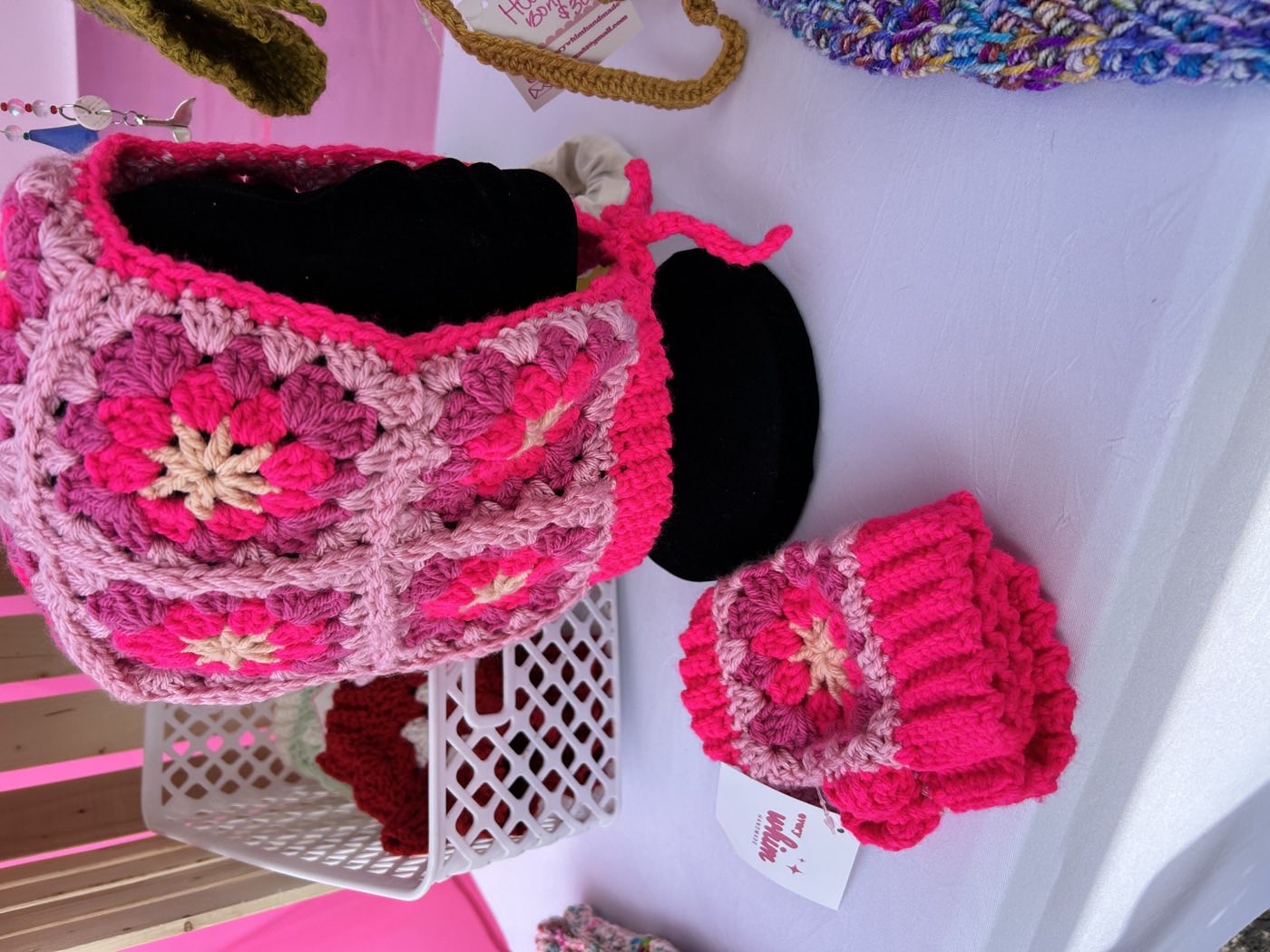 A hot pink crochet bonnet made of granny squares with cream flower centres, displayed on a stand with a matching pair of fingerless gloves in front.