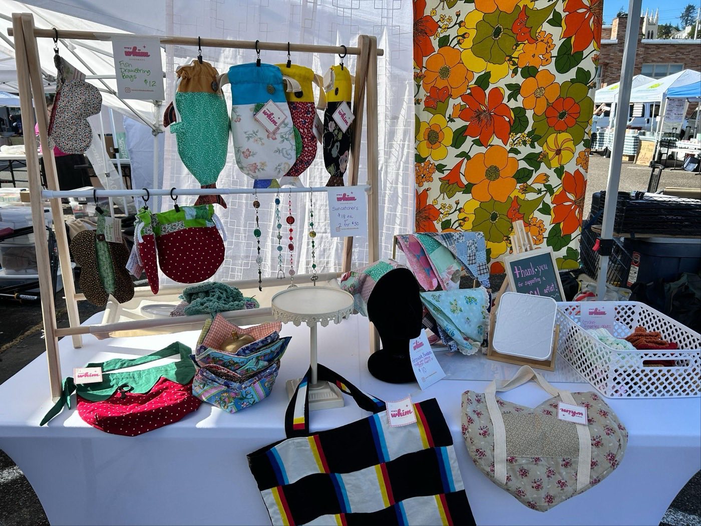 The Every Whim Handmade booth at an outdoor craft fair: fish drawstring bags hanging on a wooden rack, patchwork bandanas, suncatchers, bowl cozies and a striped patchwork tote laid out on a white table, with a bold orange and yellow vintage floral tapestry behind.