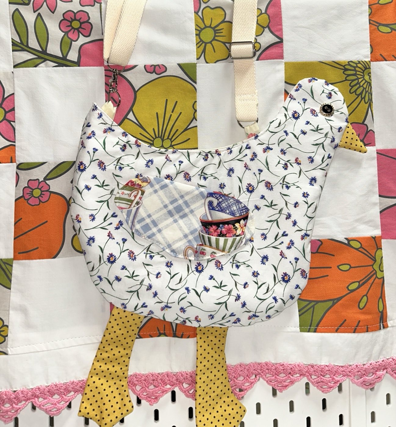 A chicken-shaped bag in white fabric printed with small blue cornflowers, with a teacup-print front pocket and yellow polka-dot legs, hung against a pink and orange patchwork quilt.