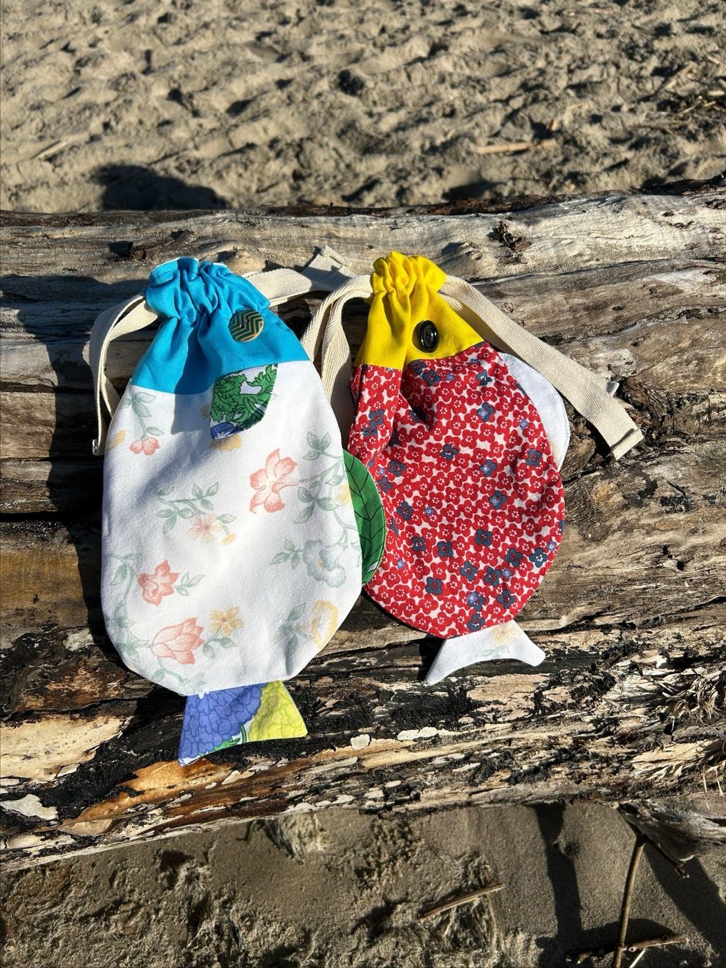 Two fish-shaped drawstring bags resting on beach driftwood: one in white floral vintage fabric with a turquoise head, the other in red flowered fabric with a yellow head, both with canvas straps.