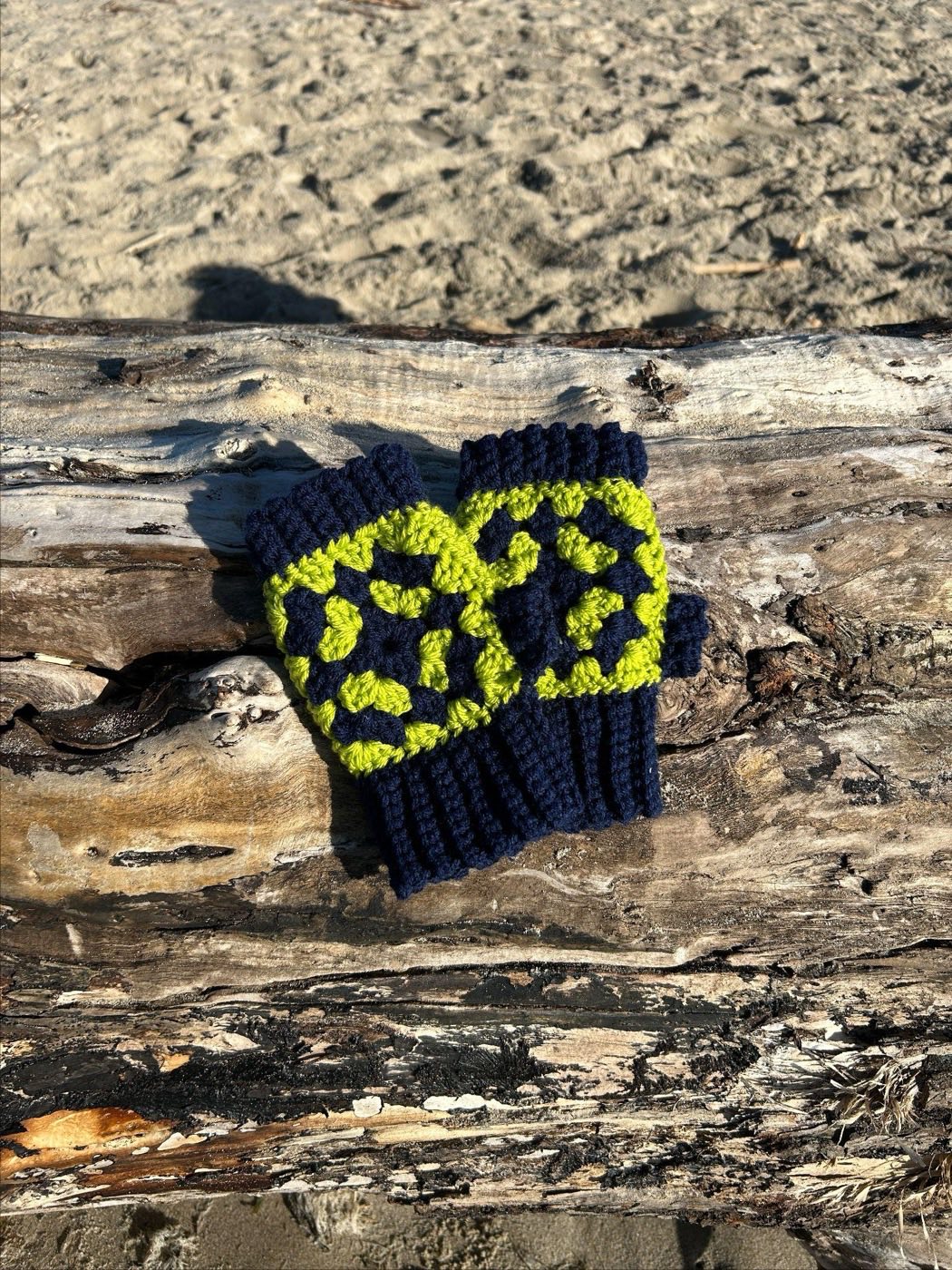 Navy blue crochet fingerless gloves with bright lime green granny squares on the back of the hand, on driftwood.