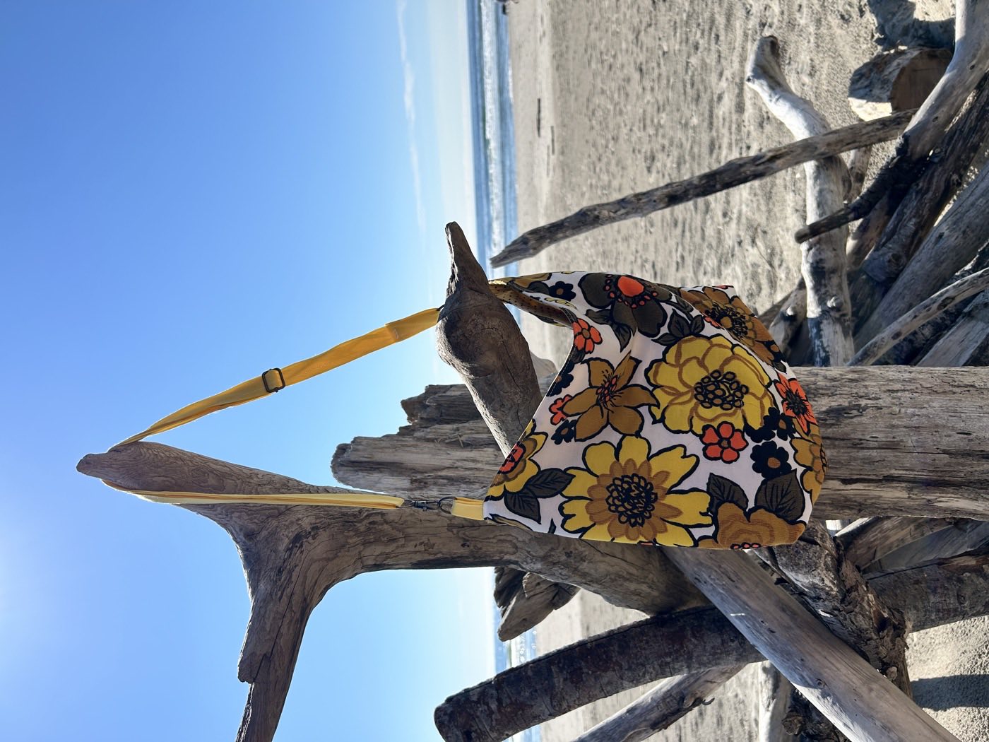 A shoulder bag in bold seventies floral fabric — mustard, orange and black blooms — with a yellow strap, hung on bleached driftwood against blue sky and open ocean.
