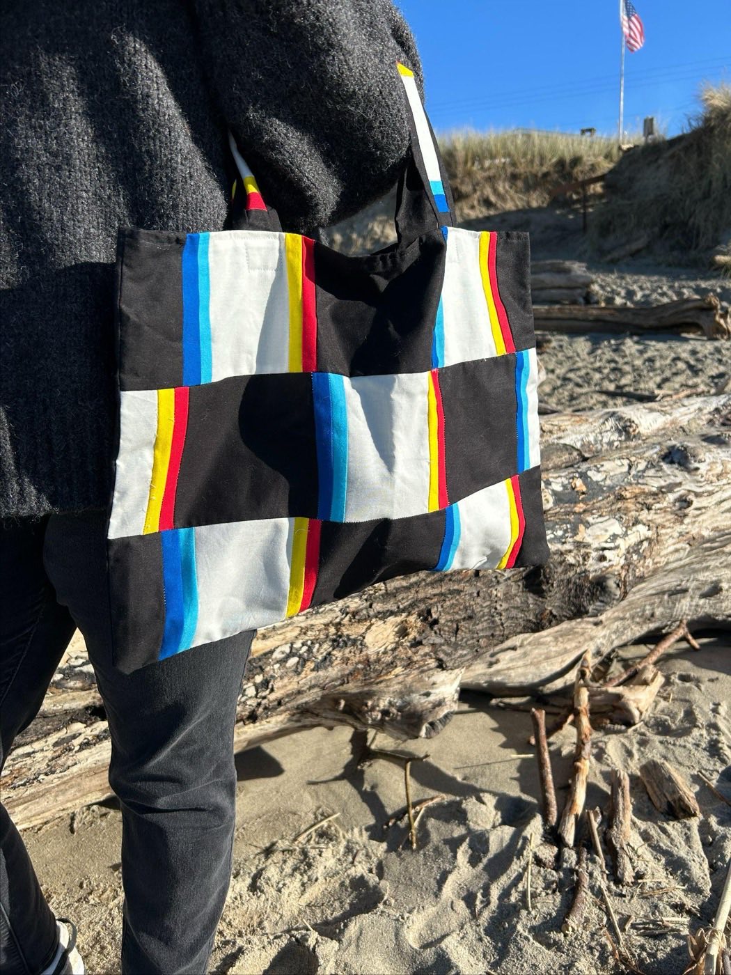 A patchwork tote bag in black and white blocks with red, yellow and blue stripes, carried on the shoulder at the beach.